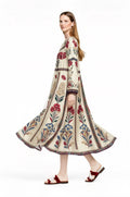 May include: A cream-colored, long-sleeve dress with a floral pattern in red, blue, and green. The dress has a flowing skirt and is worn with red sandals.