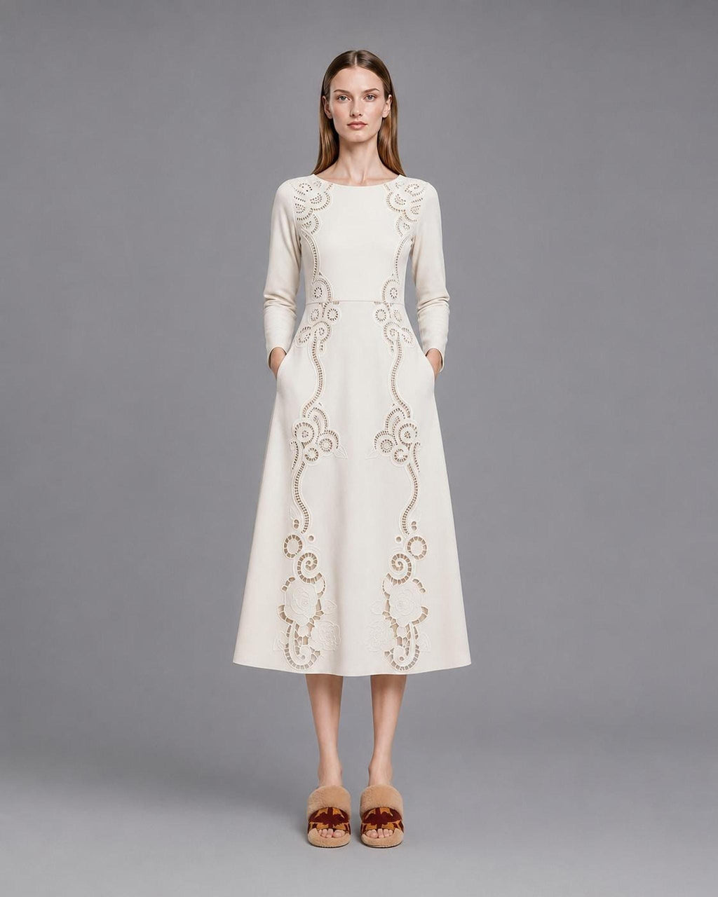 A model standing front-facing on a grey studio backdrop in a cream midi dress with long sleeves, side pockets, and intricate openwork floral scroll designs traveling from the shoulders down the skirt.
