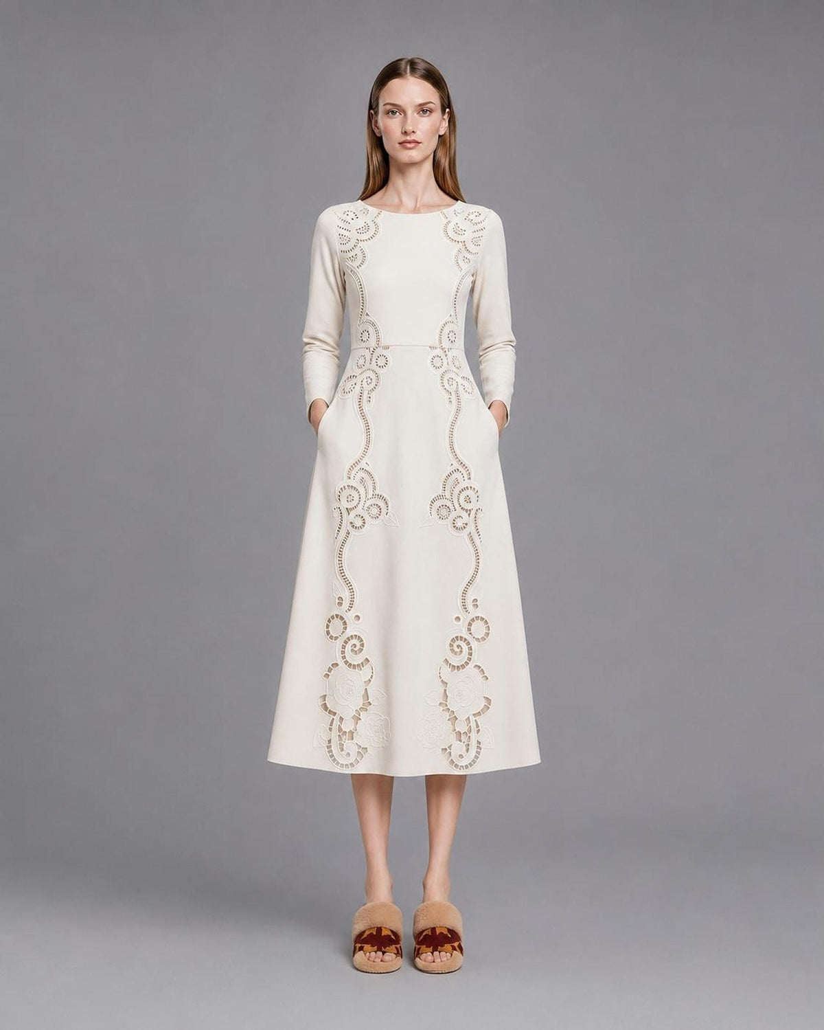 A model standing front-facing on a grey studio backdrop in a cream midi dress with long sleeves, side pockets, and intricate openwork floral scroll designs traveling from the shoulders down the skirt.
