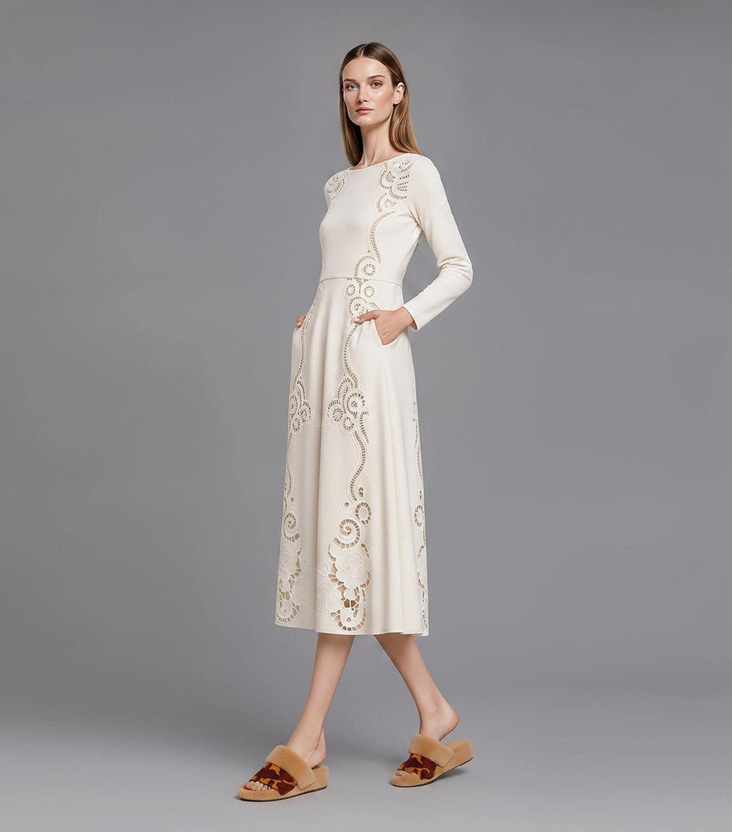 A model captured mid-stride from the right side, wearing a long-sleeved ivory dress with laser-cut floral patterns and pockets, standing against a neutral grey wall.