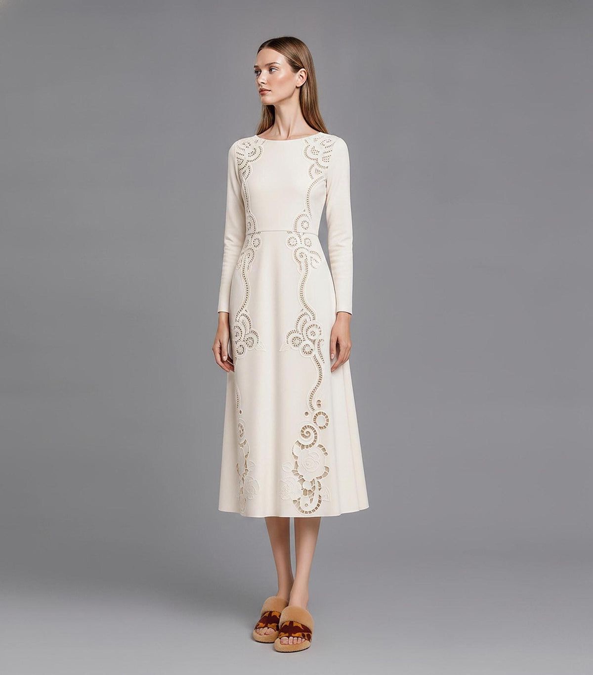 A front view of a model standing on a grey background, wearing an ivory long-sleeve A-line dress with symmetrical laser-cut scrollwork on the bodice and hem.