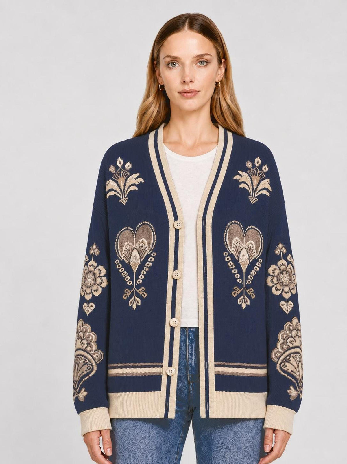A front view of a woman modeling a dark blue knit cardigan featuring beige floral and heart jacquard patterns, worn with a white tee and jeans.