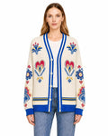 A model wearing an ivory knit cardigan with bright blue trim and colorful folk-inspired floral and heart patterns.