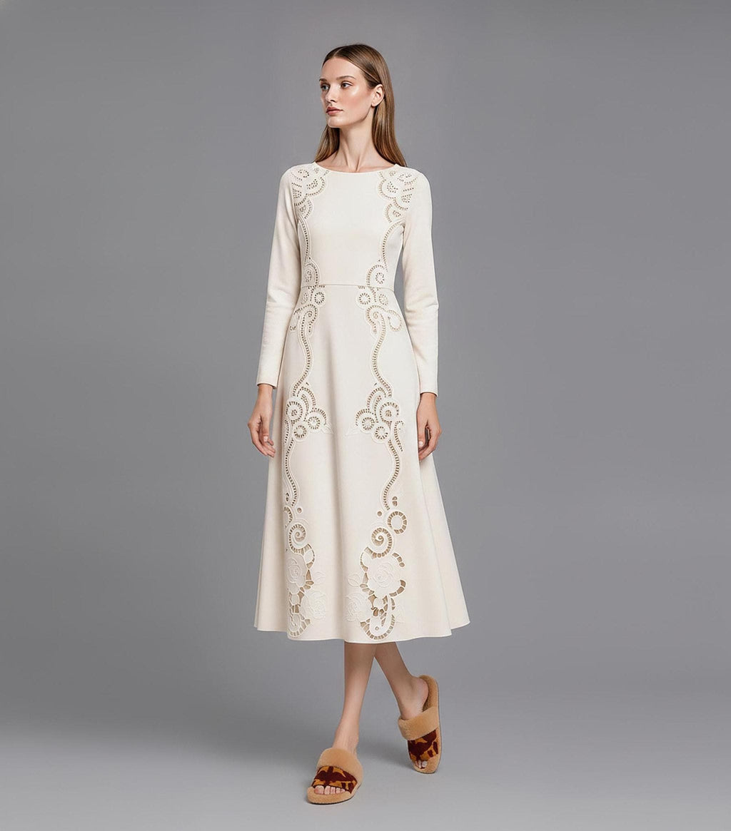 A model standing three-quarters left on a grey studio background wearing a long-sleeve ivory midi dress with laser-cut filigree scrollwork down the front and brown fuzzy slide sandals.