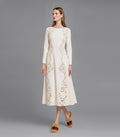 A model standing three-quarters left on a grey studio background wearing a long-sleeve ivory midi dress with laser-cut filigree scrollwork down the front and brown fuzzy slide sandals.