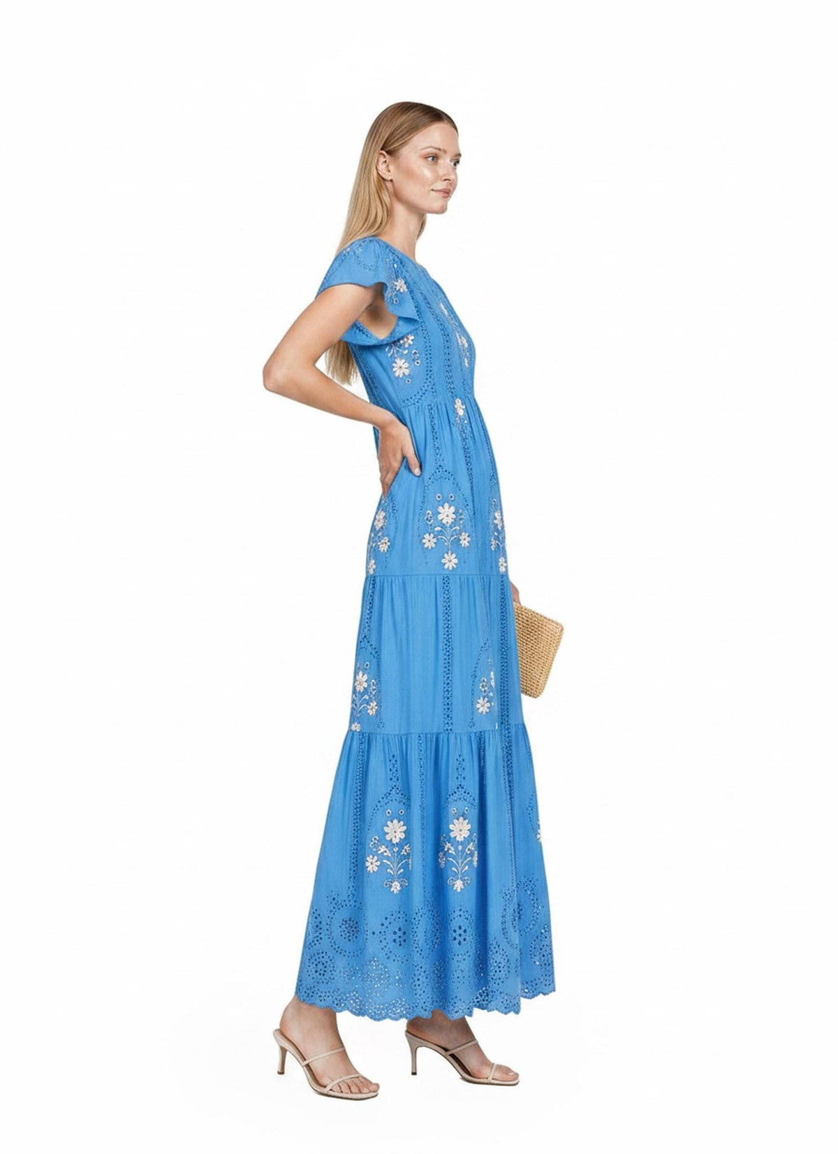 A full-length side shot of a model standing to the left, wearing a blue tiered maxi dress with white floral embroidery while holding a straw handbag.