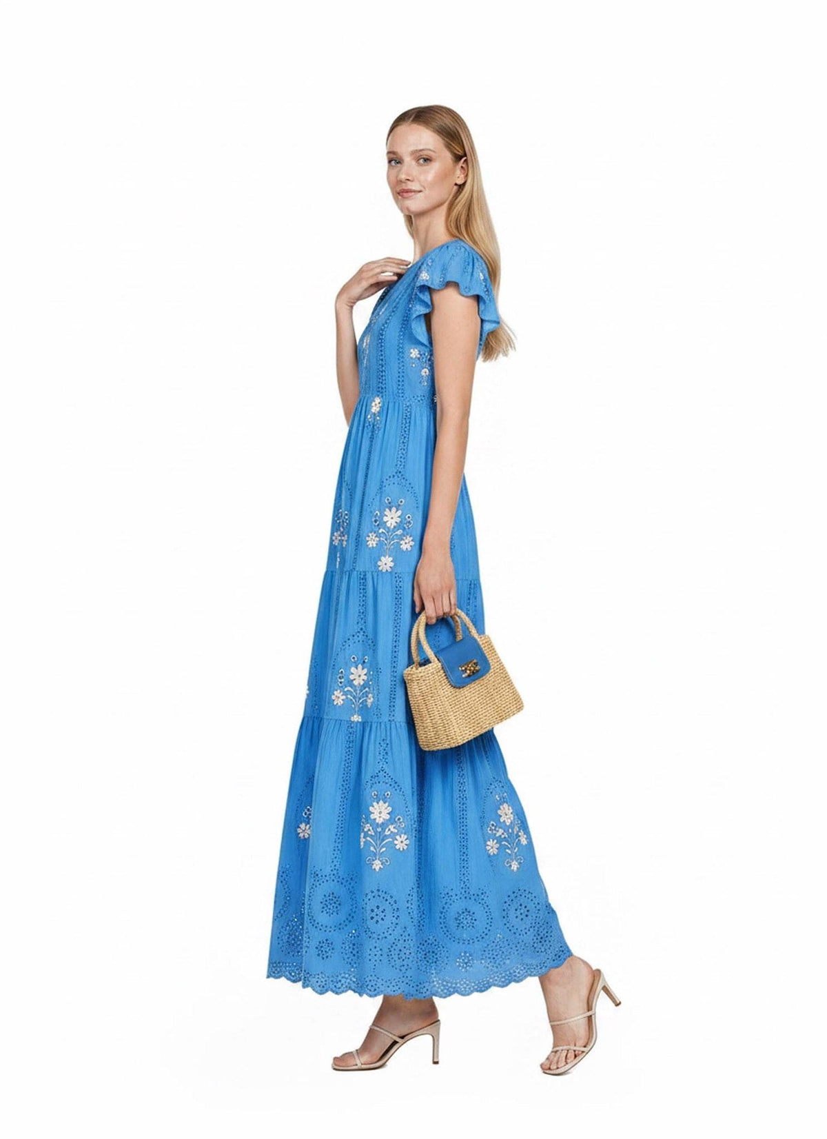 A model posing in profile holding a small straw handbag with a blue flap, wearing a blue tiered maxi dress with eyelet and floral embroidery details.