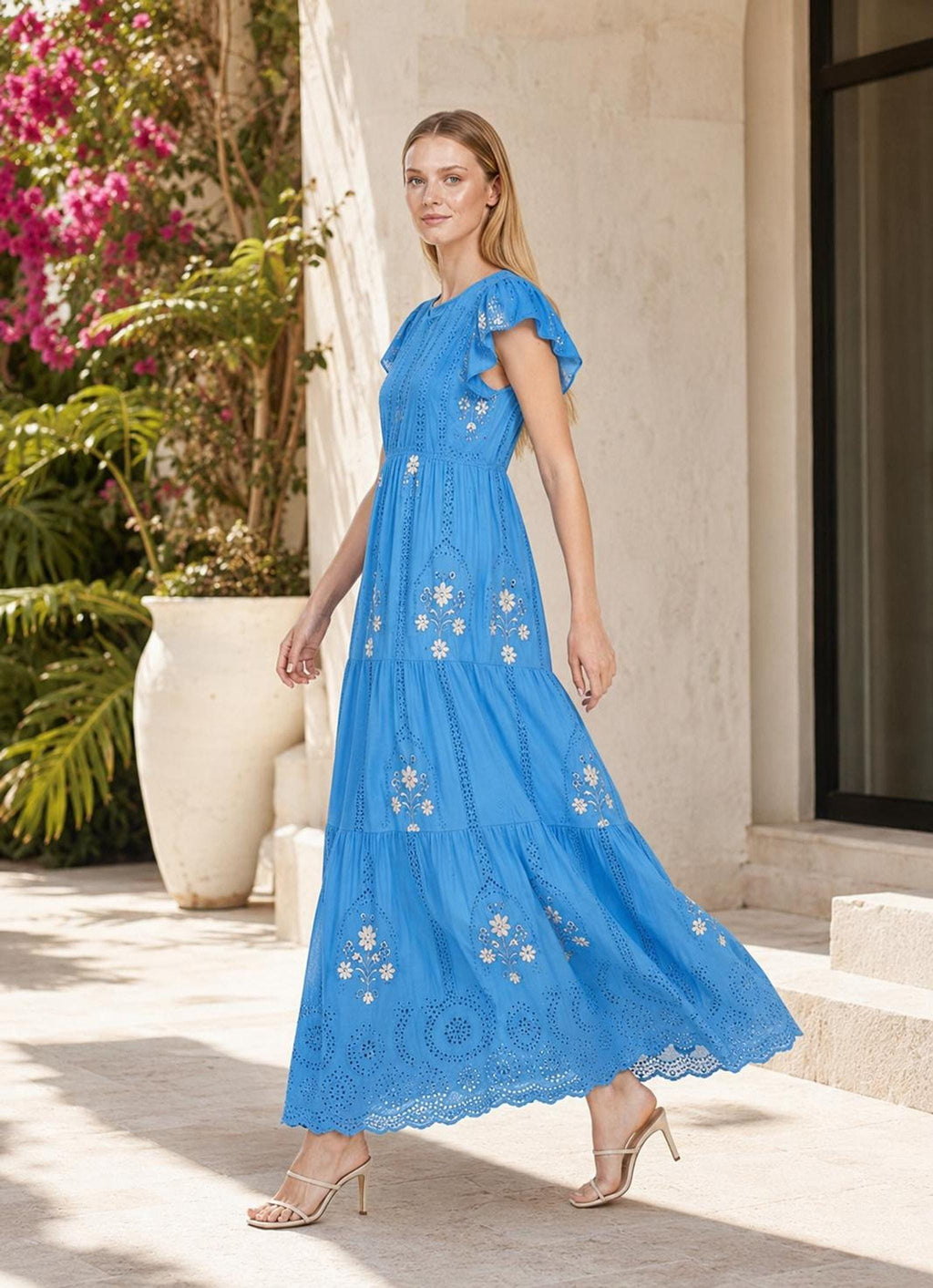 An outdoor full-length photo of a model walking near a stone wall, wearing a bright blue tiered maxi dress with floral embroidery.