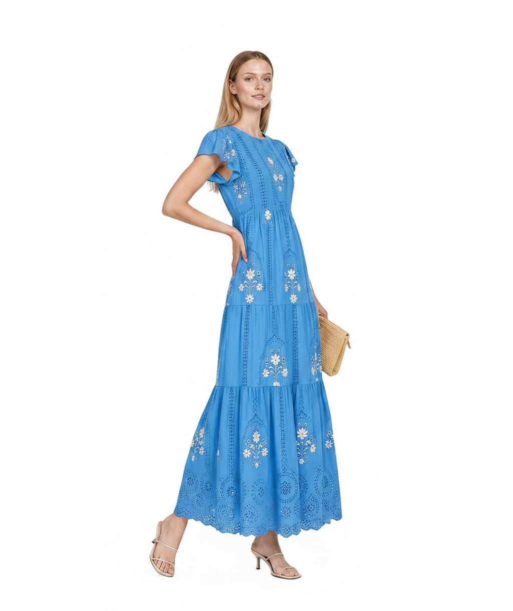 A model standing three-quarters left wears a bright blue tiered maxi dress with flutter sleeves and white floral embroidery, holding a small straw clutch.