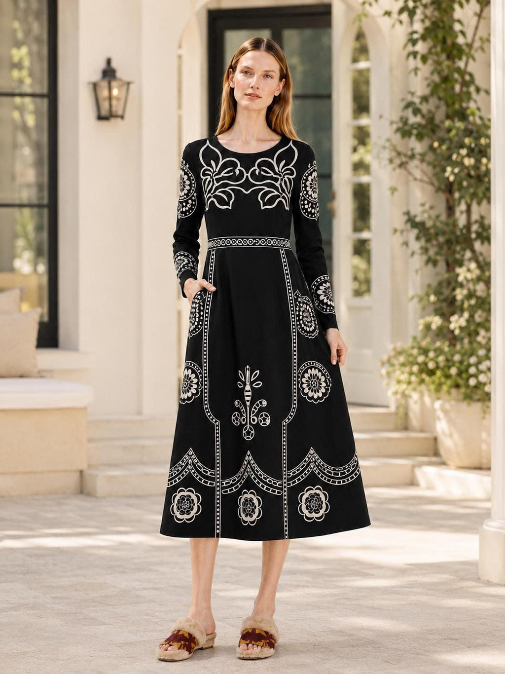 A model poses outdoors near a white building and potted plants, wearing a black long-sleeve midi dress with beige folk floral details.