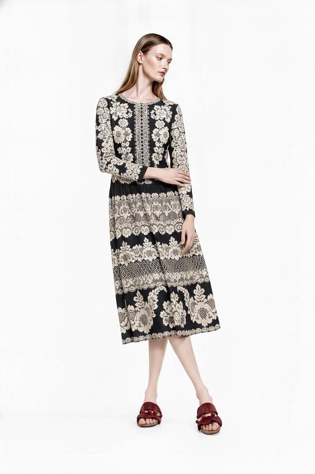 May include: A long-sleeved midi dress featuring a black base with a repeating cream-colored floral and lace-inspired pattern. The garment has a round neckline, a fitted bodice, and a gathered skirt that falls below the knees, paired with dark red slides.