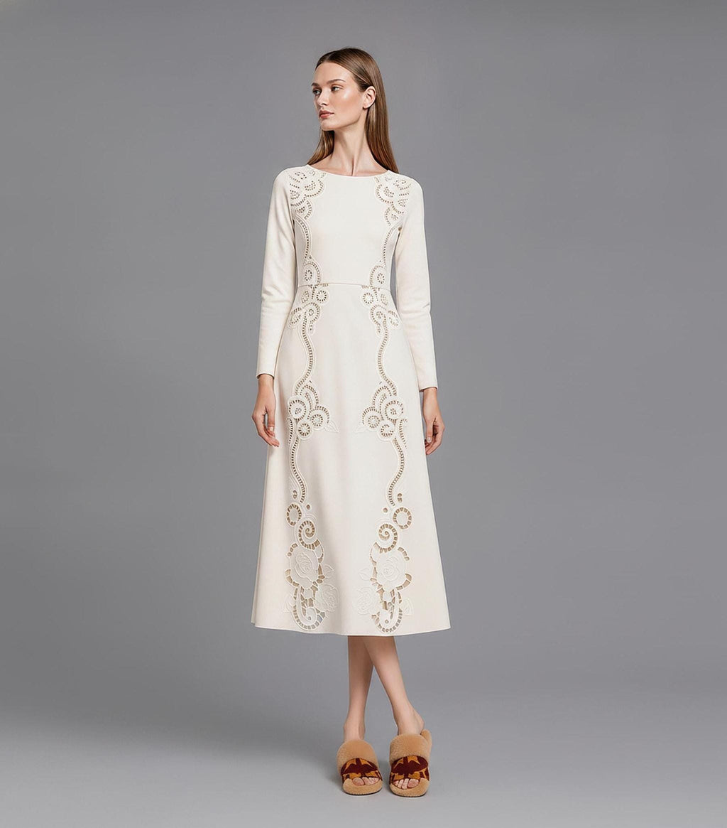 A model facing slightly left on a grey studio background in a long-sleeve ivory A-line midi dress featuring ornate cutout patterns along the neckline and skirt, paired with brown fur slides.