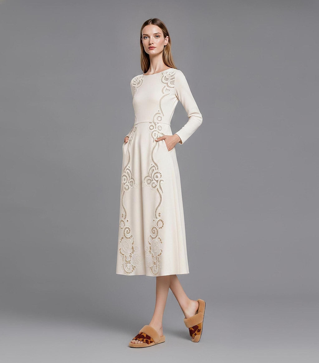 A full-length view of a model in profile against a grey backdrop, showcasing an ivory long-sleeved midi dress with decorative laser-cut embroidery and side seam pockets.