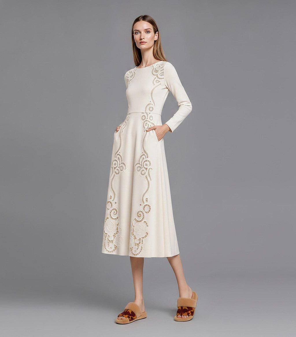 A model standing at a slight angle on a grey studio floor, wearing a cream-colored long-sleeve midi dress adorned with intricate cutout scroll and rose designs, with her hands resting in the dress pockets.