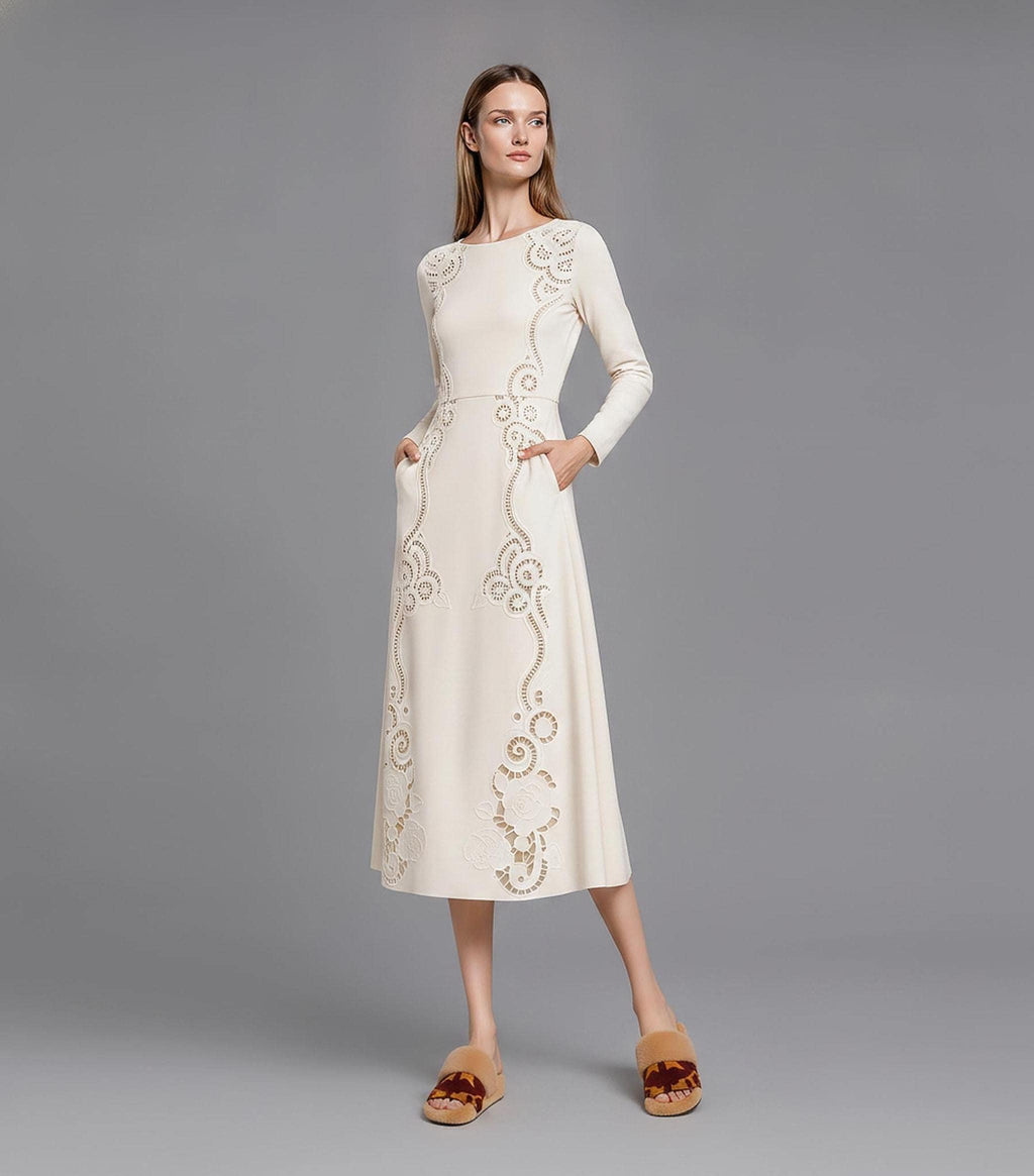A model posing upright on a grey studio background in an ivory long-sleeve dress with cutout lace-like floral panels sweeping down the bodice and flared skirt.