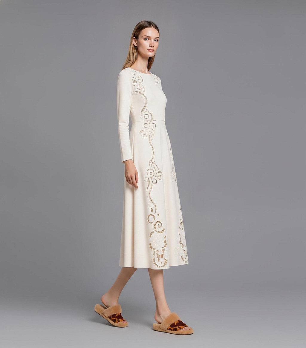 A side-profile view of a model walking across a grey studio floor in a long-sleeve cream midi dress featuring elaborate cutout embroidery patterns.