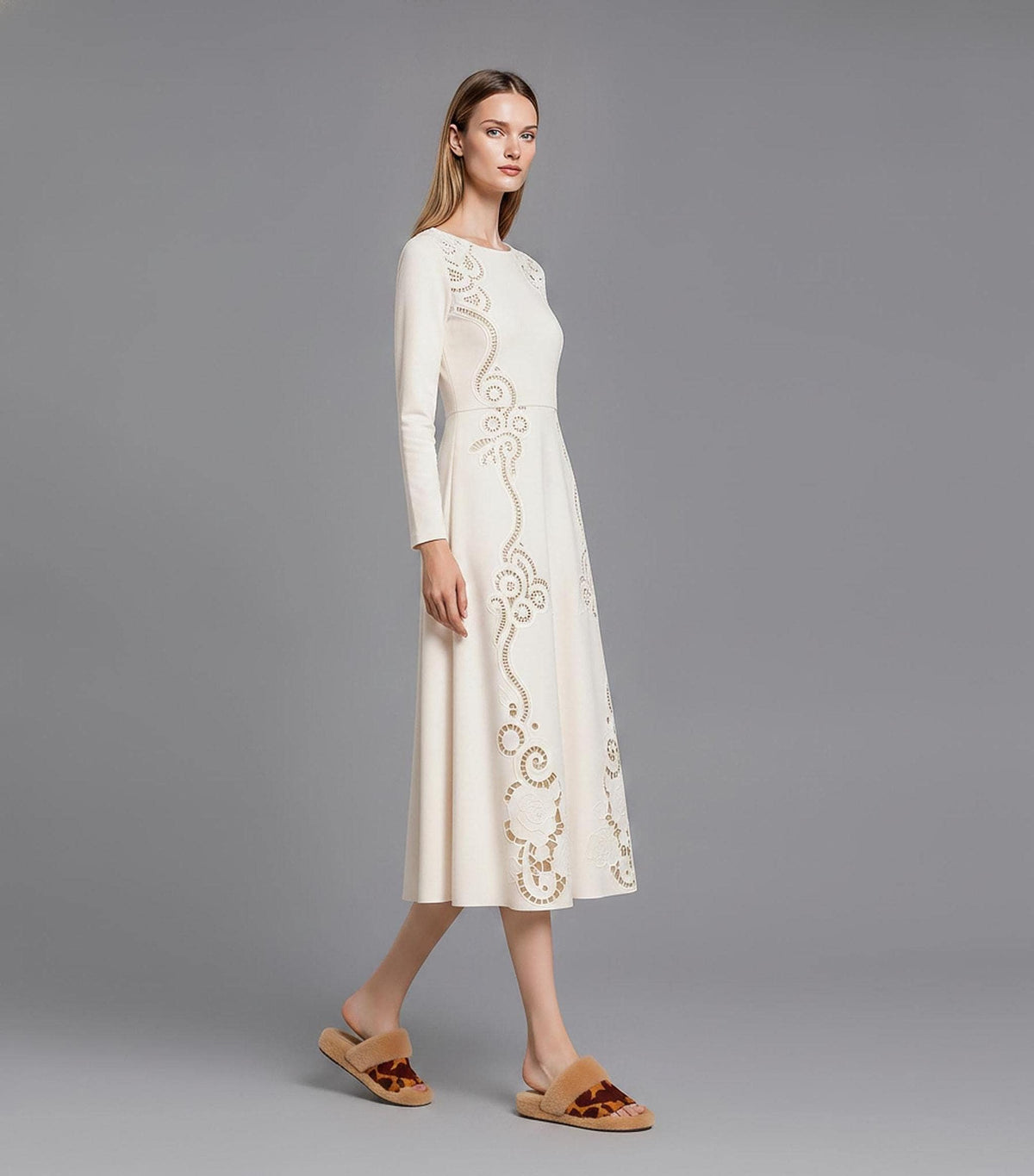 A side-profile view of a model walking across a grey studio floor in a long-sleeve cream midi dress featuring elaborate cutout embroidery patterns.