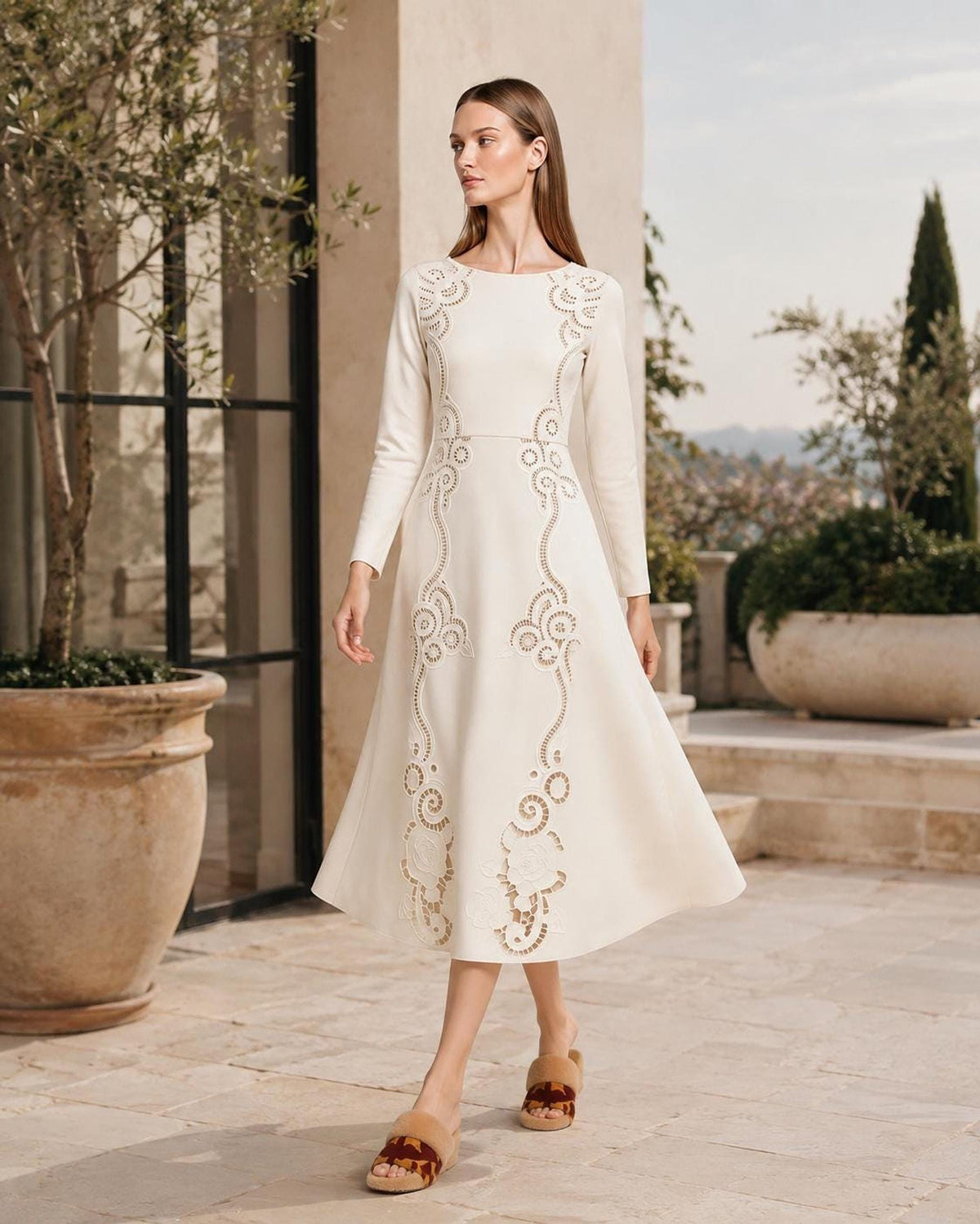 A model walking outdoors on a paved patio in an ivory long-sleeve midi dress featuring intricate cutout patterns, with large potted plants and a building facade in the background.