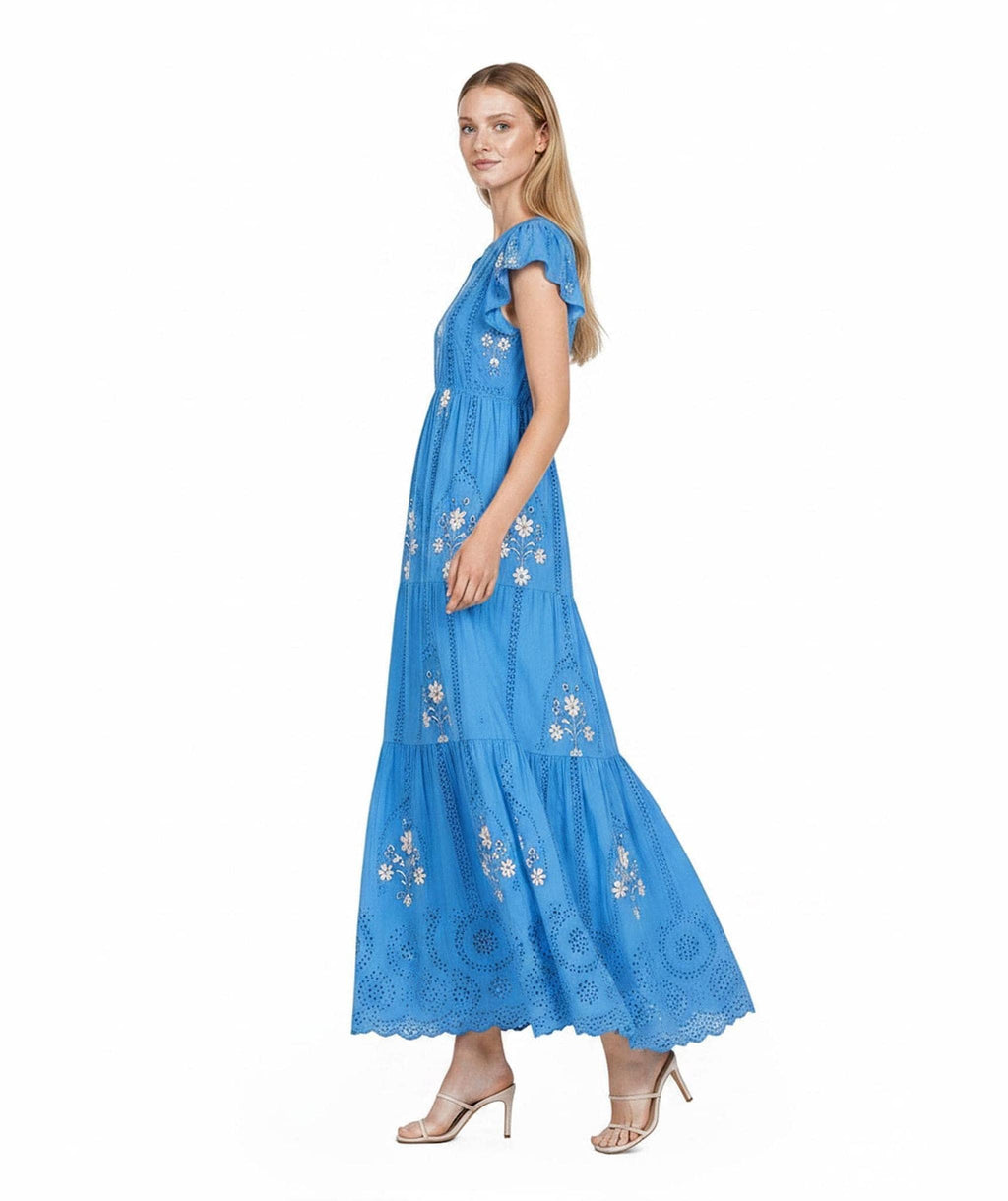 A full-length side profile view of a model in a bright blue tiered maxi dress with floral embroidery and cutouts, standing on a white background.