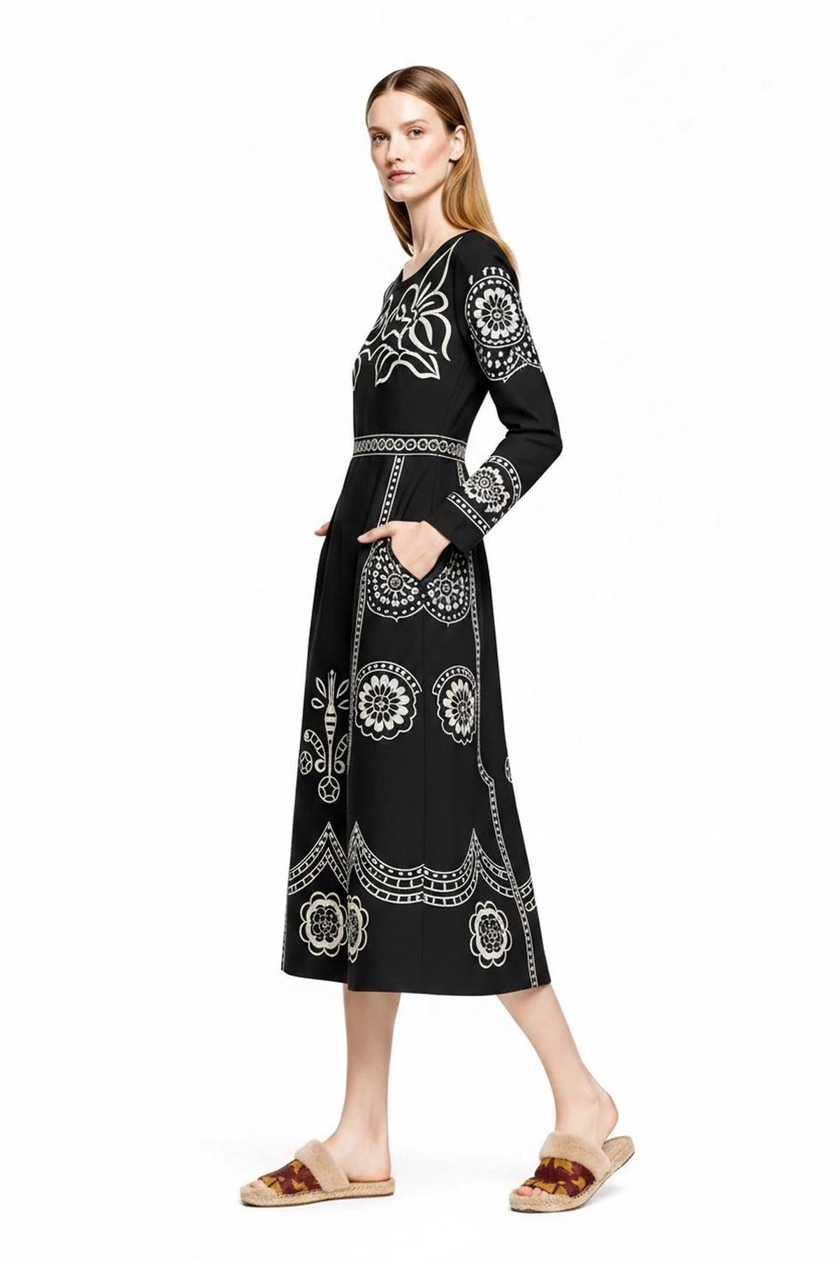 A model stands in profile against a plain white background, wearing a black long-sleeve midi dress with beige folk floral embroidery and fuzzy slip-on sandals.