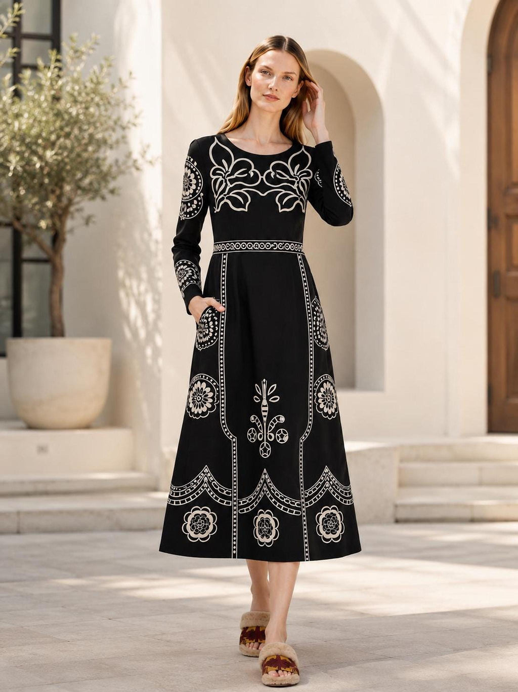 A model stands outdoors on a paved patio in front of a building, wearing a black long-sleeve midi dress with beige folk floral prints and fuzzy slippers.