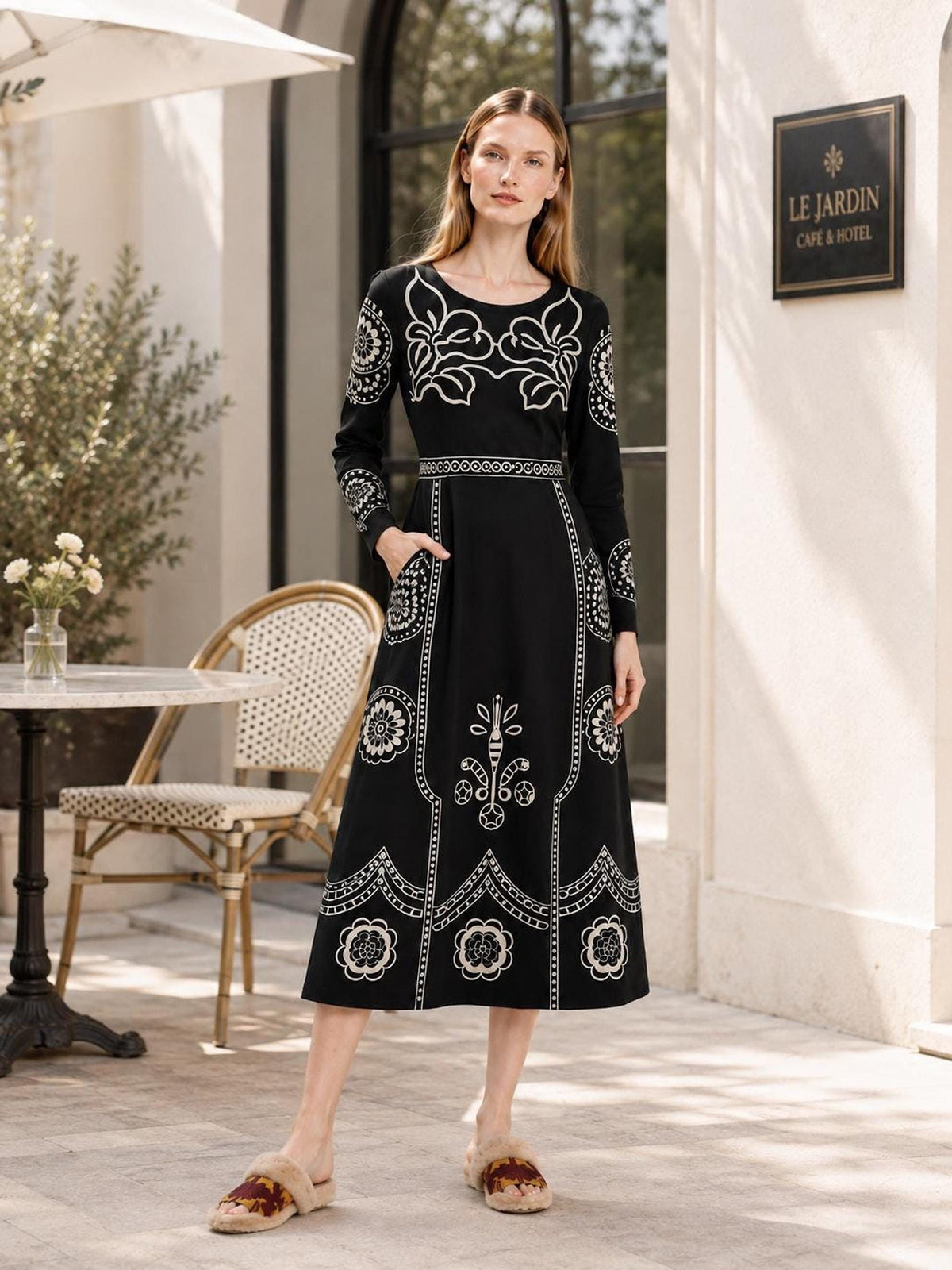 A model stands on an outdoor patio near a cafe table and chairs, wearing a black long-sleeve midi dress adorned with beige folk floral patterns.