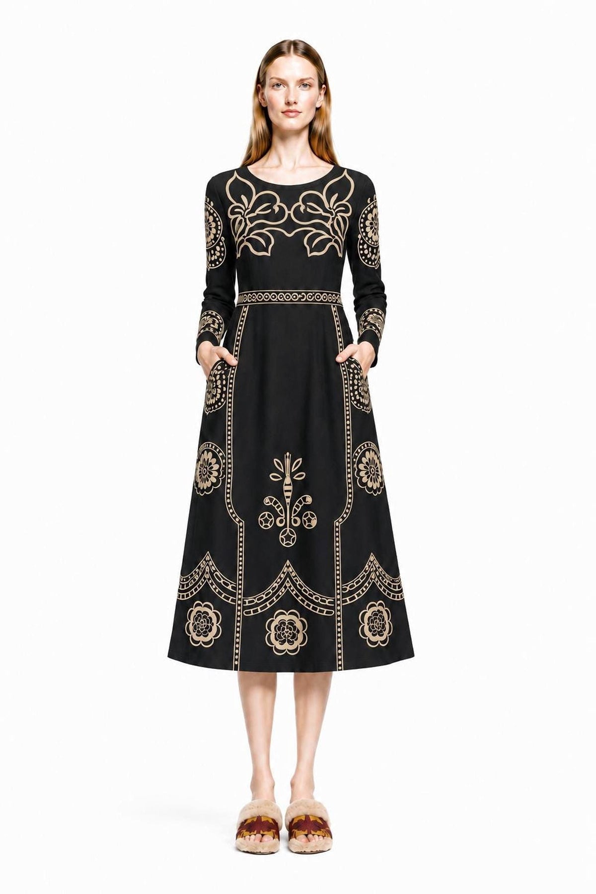 A model stands facing forward wearing a black midi dress with beige folk floral print and long sleeves against a plain white background.